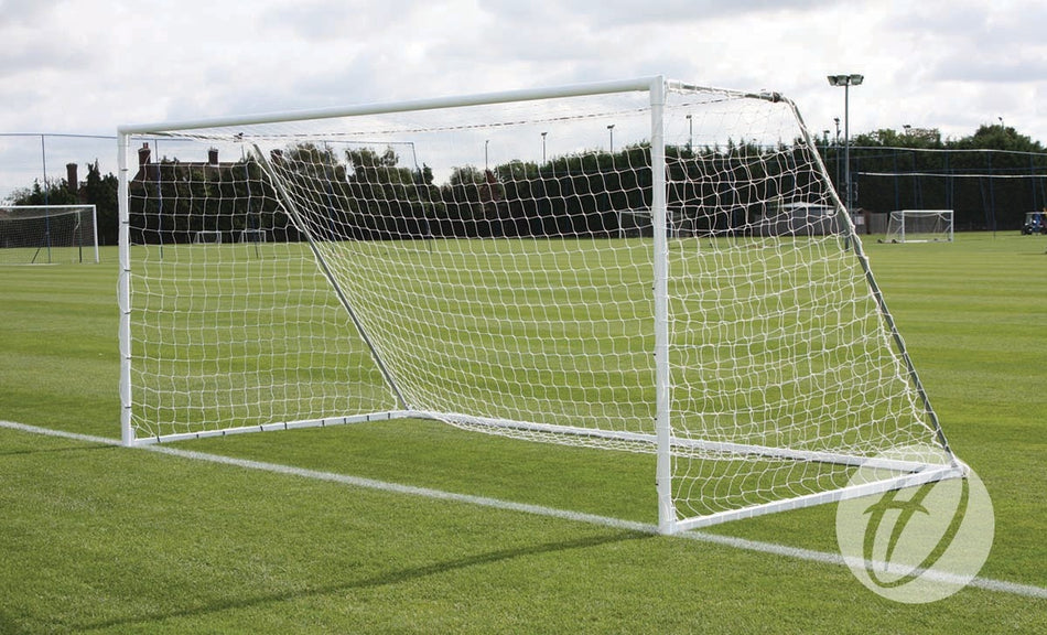 A Harrod Sport Heavyweight Freestanding Football Goal - 9v9 with a white frame and net is positioned on a well-maintained grass field under a cloudy sky, with trees and buildings visible in the background.