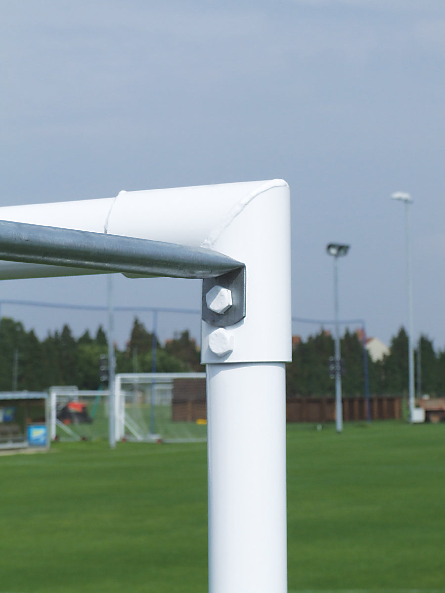 Close-up of the top corner of a Harrod Sport soccer goalpost, equipped with Continental Tubular Steel Net Supports - Senior/Junior, constructed from zinc plated steel with visible bolts. The white metal goal is set on a green grass field, and other full size goals and trees appear blurred in the background beneath a blue sky.