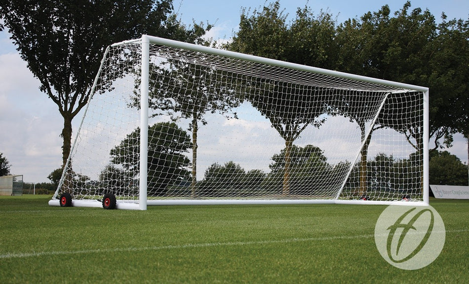 A white aluminium Harrod Sport 4G Portagoal - Junior with a net and wheels stands on green grass, with trees and a partly cloudy sky in the background.