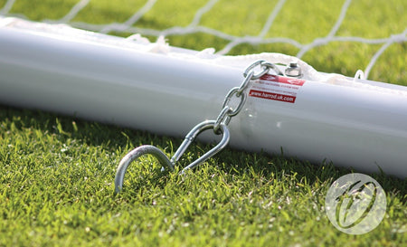 A close-up of the Harrod Sport 4G Portagoal - Junior, an aluminium football goalpost secured to grass with a chain and hook, with part of the net visible in the background.