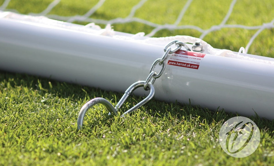 A close-up of the Harrod Sport 4G Portagoal - Junior, an aluminium football goalpost secured to grass with a chain and hook, with part of the net visible in the background.