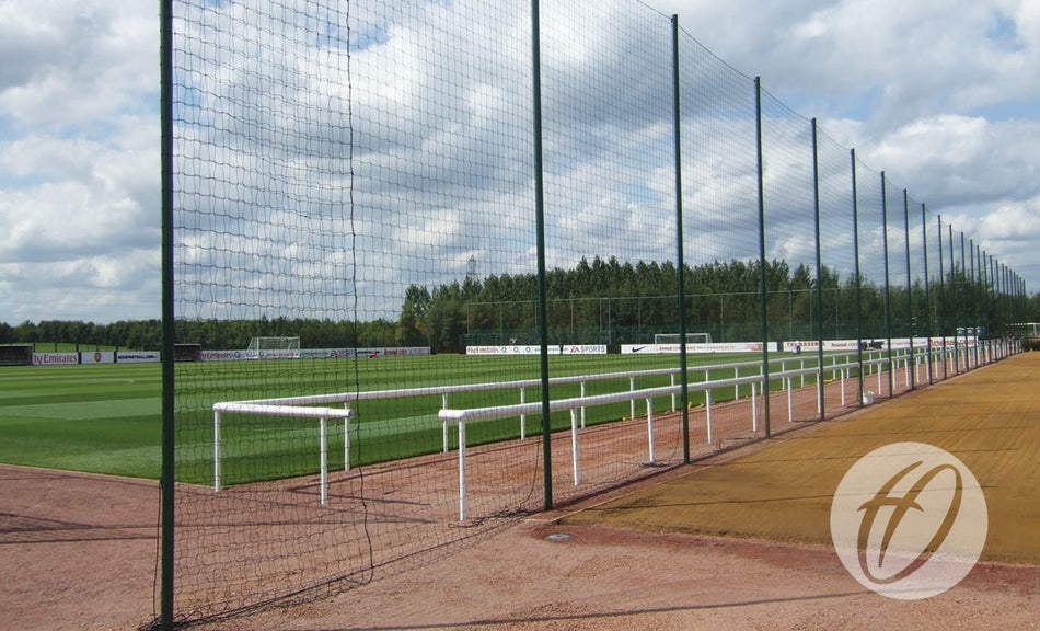 A fenced soccer training ground with green grass, white goalposts, ball stop netting, and a dirt track, bordered by Harrod Sport 6.4m high Perimeter Poles. Trees and advertisement boards line the background under a partly cloudy sky.