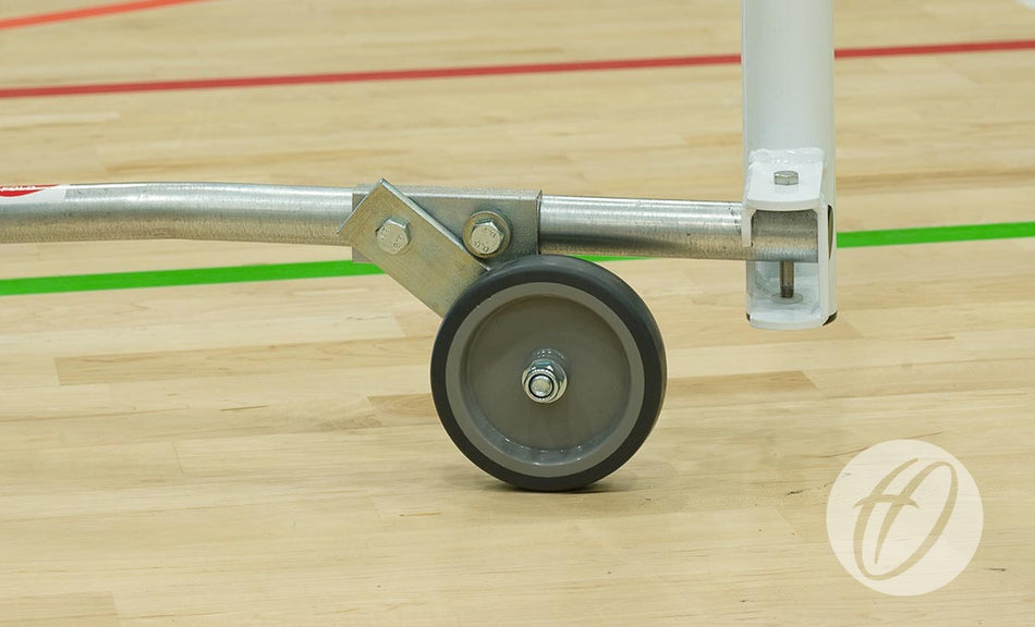 A close-up of a gray caster wheel attached to the folding side frame of the Harrod Sport FS8 Folding Wheelaway Goal (4.88m x 1.22m), positioned on a wooden gym floor marked with red and green lines.