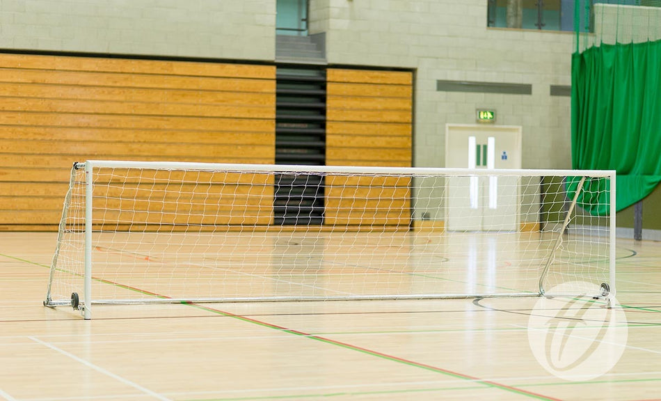 An indoor sports hall with a Harrod Sport FS8 Folding Wheelaway Goal (4.88m x 1.22m) in the foreground, featuring a folding side frame design, wooden bleachers in the background, and green curtains hanging on the right side.