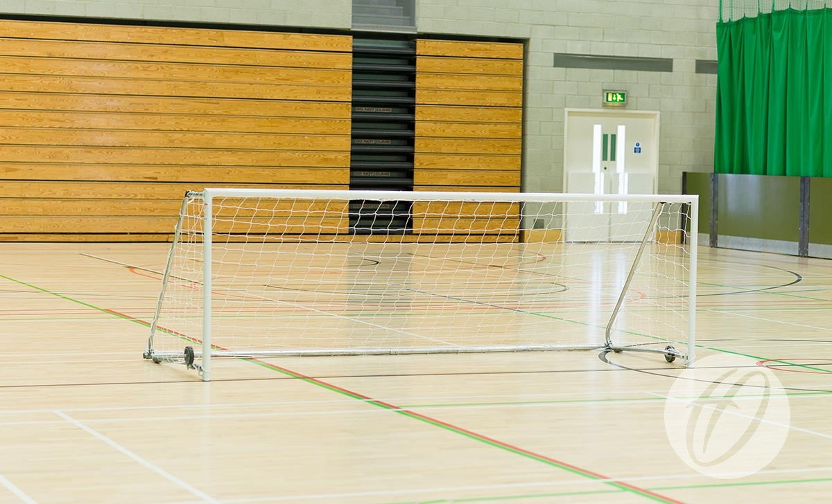 A Harrod Sport FS8 Folding Wheelaway Goal (3.66m x 1.22m) stands on an empty gym court with wooden bleachers, green curtains, and a white double door in the background.