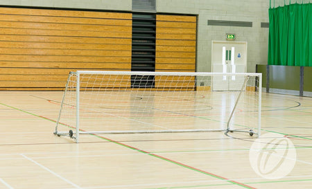 A Harrod Sport FS8 Folding Wheelaway Goal (3.66m x 1.22m) stands on an empty gym court with wooden bleachers, green curtains, and a white double door in the background.