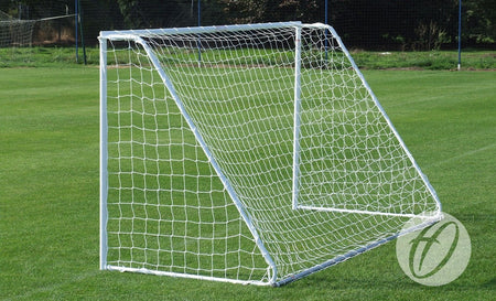 A Harrod Sport Freestanding Steel Football Goal - 7v7/5v5, featuring a compact white frame and a net, is positioned on a well-maintained grass field, with a fence and greenery visible in the background.
