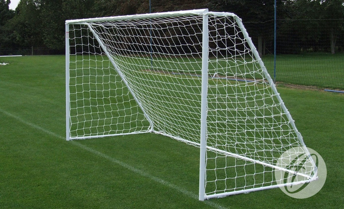 A Harrod Sport Freestanding Steel Football Goal, measuring 16′x6′, with galvanised steel back supports and a net is positioned on a green grass field. Trees and fencing can be seen in the background.