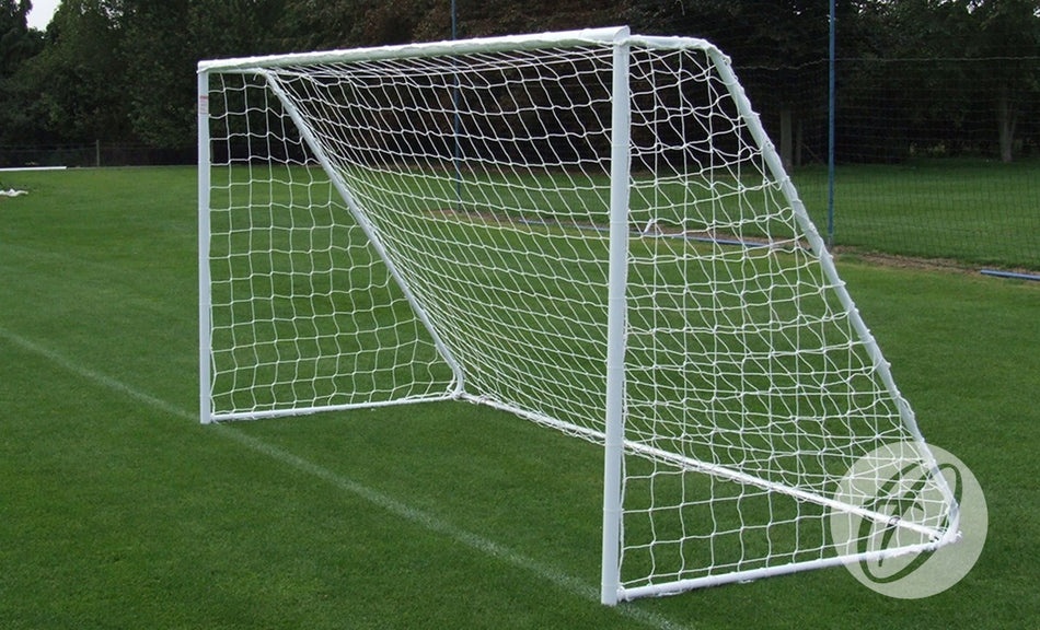 A Harrod Sport Freestanding Steel Football Goal, measuring 16′x6′, with galvanised steel back supports and a net is positioned on a green grass field. Trees and fencing can be seen in the background.