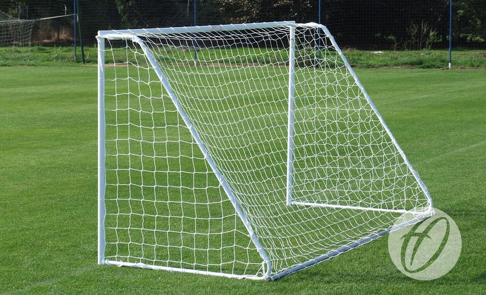 A Harrod Sport Freestanding Steel Football Goal - 16′x6′, complete with a net and galvanised steel back supports, stands unused on a well-maintained green grass field. In the background, there is more grass, along with a fence and trees.