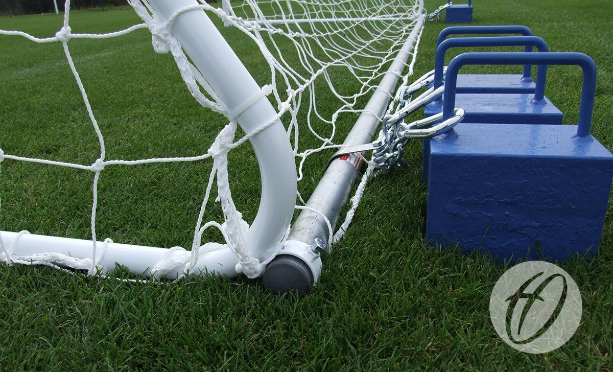 A close-up of a Harrod Sport Freestanding Steel Football Goal – 16′x6′, secured on a green grass field with metal chains and blue weights. The net is attached to the freestanding, galvanised steel goalpost with robust steel back supports, while part of a circular logo can be seen in the corner.