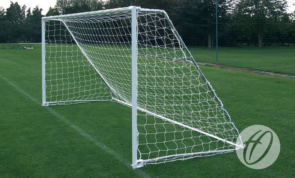 A Harrod Sport Folding Steel Football Goal - 16′x6′, complete with a net and convenient folding side frame, is set up on a green grass field with trees and a fence visible in the background.