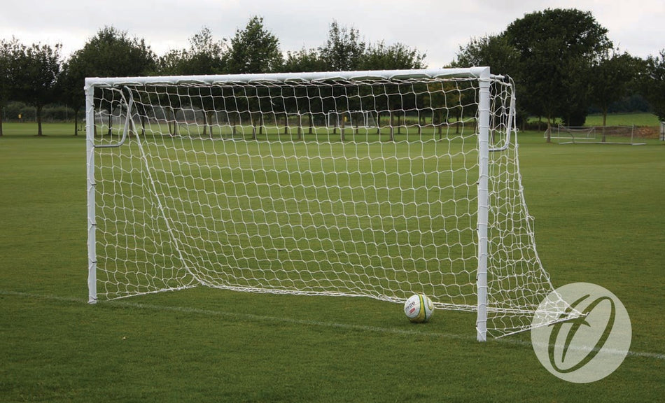 A Harrod Sport Heavyweight Football Goal - 16′x6′ stands on a grassy field with a soccer ball resting just inside the net. Trees and a cloudy sky form the background, enhancing the scene around this sturdy, heavy duty football goal.
