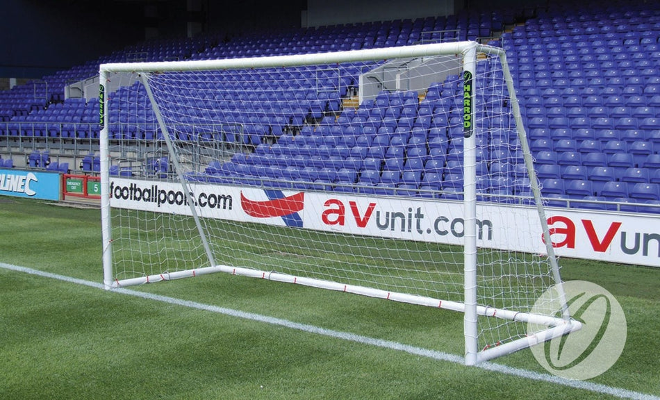 A Harrod Sport Polygoal - 7v7/5v5 freestanding white PVC football goal with net is positioned on a green grass field in front of empty blue stadium seats, with advertising boards visible behind the lightweight goal.