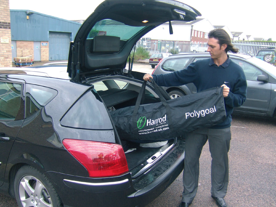 A man places a black Harrod Sport Polygoal - 7v7/5v5 bag, designed for transporting a portable soccer goal, into the trunk of a black car parked outdoors on a cloudy day near some buildings and other vehicles.