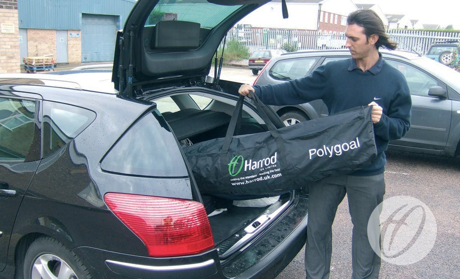 A man stands by an open car trunk in a parking lot, holding a large black Spare Polygoal Bag from Harrod Sport—possibly bringing it for replacement on a cloudy day.