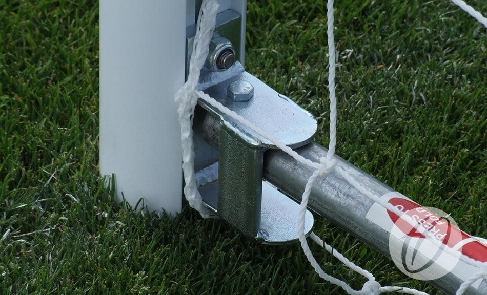 Close-up of a metal hinge and bolt connecting a metal rod to a white pole, with a net attached, on a grassy field. The image highlights the sturdy construction characteristic of the Harrod Sport Folding Aluminium Football Goal - 7v7/5v5, with part of the distinctive red and white Harrod Sport label visible on the rod.