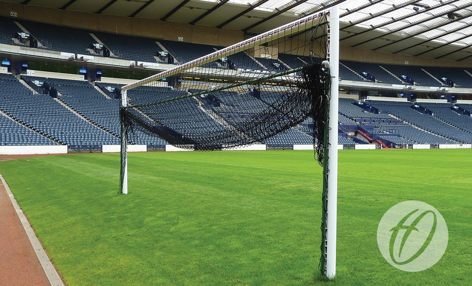 A Harrod Sport Hinged 3G Aluminium Bottom Net Support - Senior goal with a tangled, sagging net stands on a green field in an empty stadium lined with blue seats.