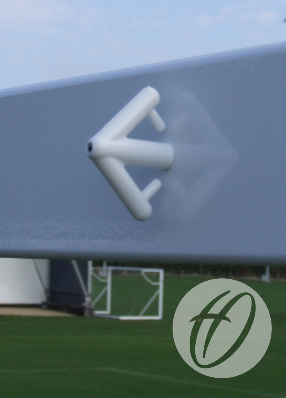 A Harrod Sport Synthetic Arrow Net Hook from the 12-pack is attached to a horizontal steel football goal post, with a blurred soccer field and goal behind. A circular logo with stylized letters appears in the lower right corner.