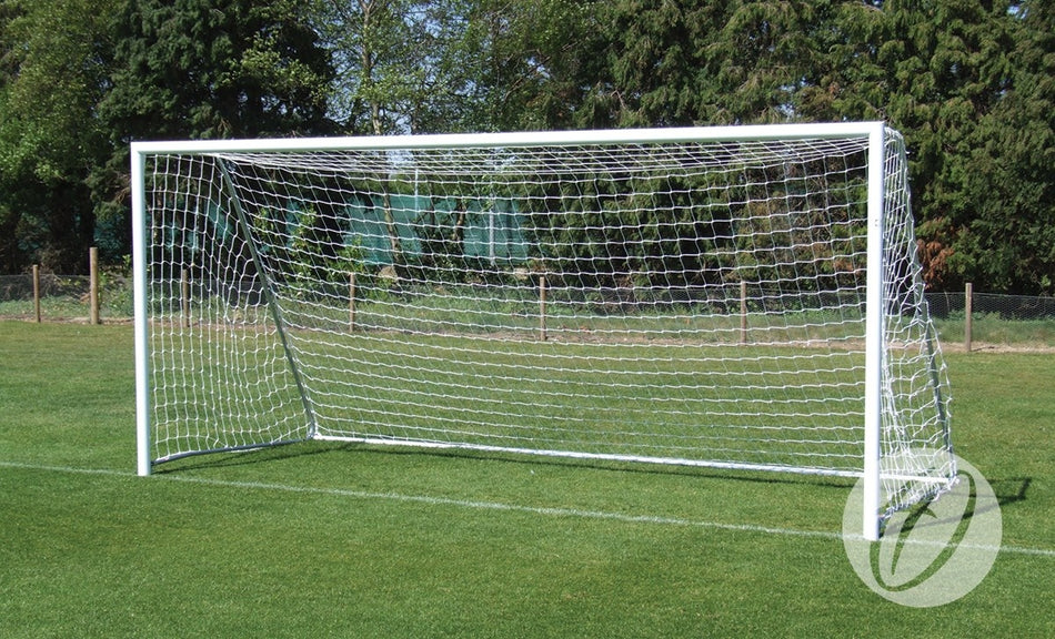 A Harrod Sport Folding Aluminium Football Goal – 9V9 with white folding side frames stands on a green grass field, surrounded by trees and a fence in the background on a sunny day.