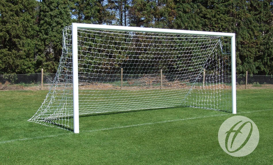 A Harrod Sport 4G Stadium Goal - 9V9 made of white aluminium with netting stands on green grass, with a fence and tall trees in the background beneath a blue sky.
