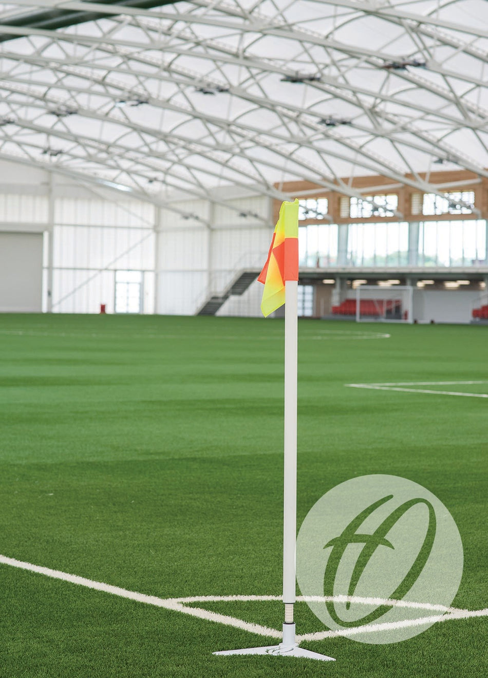 A Harrod Sport White Pro Flex 50mm Pole with Flag, Socket & Lid stands on an indoor artificial turf soccer field marked with white lines. The facility features a curved white roof and empty red seating in the background, providing injury reduction features that meet professional club flagpole standards.