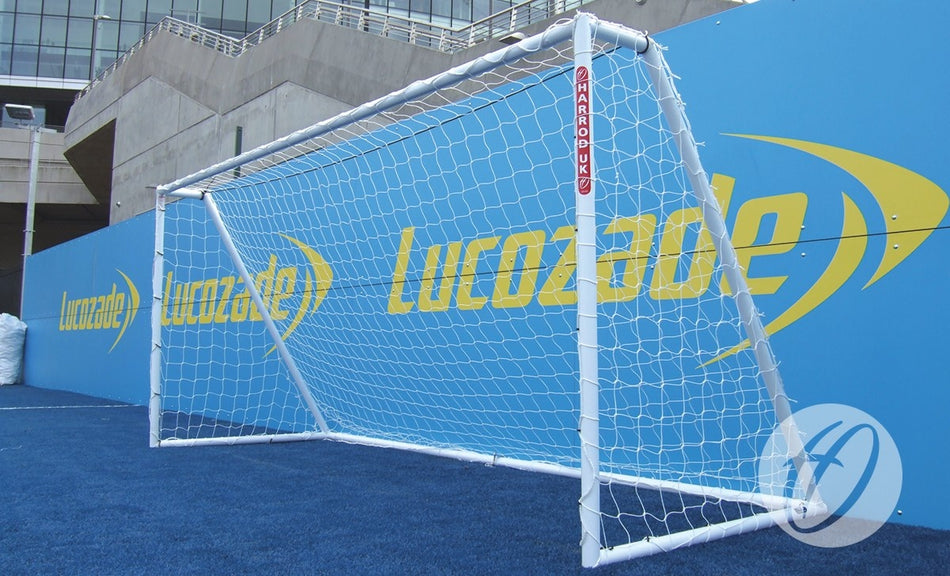 A Harrod Sport Heavy Duty Galvanised Goal (3.66m x 1.83m - 7V7/5V5) stands on blue artificial turf in front of a large blue wall featuring yellow Lucozade logos. The weather appears clear, and a stadium building is visible in the background, emphasizing the robust welded side frames characteristic of this heavy-duty football goal model.