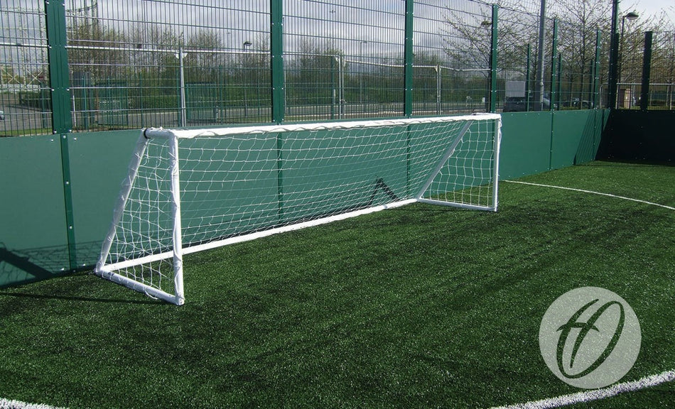 A Harrod Sport Heavy Duty Galvanised Goal, measuring 4.88M x 1.22M (5V5), stands on a green artificial turf field enclosed by high metal fencing. Built from durable galvanised steel with a welded side frame, this football goal is designed for longevity, with trees and a parking lot visible in the background.