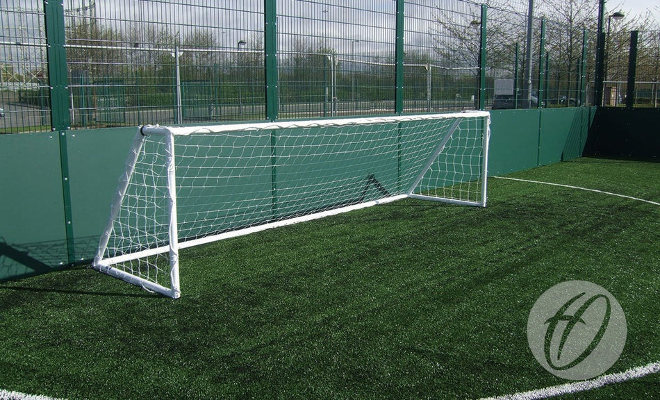 A Harrod Sport Heavy Duty Galvanised Goal, measuring 3.66m x 1.22m (5V5), made from galvanised steel with a welded side frame, is positioned on artificial turf inside a fenced outdoor field. In the background, trees and a parking lot are visible, emphasizing the goal's robust construction and suitability for demanding environments.