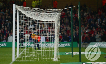 A football goal on the pitch features a damaged, sagging net due to the absence of the Harrod Sport Freehanging Net Support System - 4 Poles. In the background, a blurred crowd and security are visible, with a logo shown in the bottom right corner of the image.
