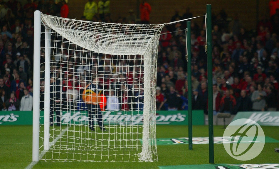 A football goal on the pitch features a damaged, sagging net due to the absence of the Harrod Sport Freehanging Net Support System - 4 Poles. In the background, a blurred crowd and security are visible, with a logo shown in the bottom right corner of the image.