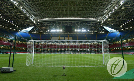 A view of an empty soccer stadium, showing the goalpost and net from behind. The stands are empty, and the stadium roof is partially open. A Harrod Sport Freehanging Net Support System - 4 Poles, designed to meet UEFA regulation net support standards, is clearly visible near the goal. A logo is visible in the lower right corner.