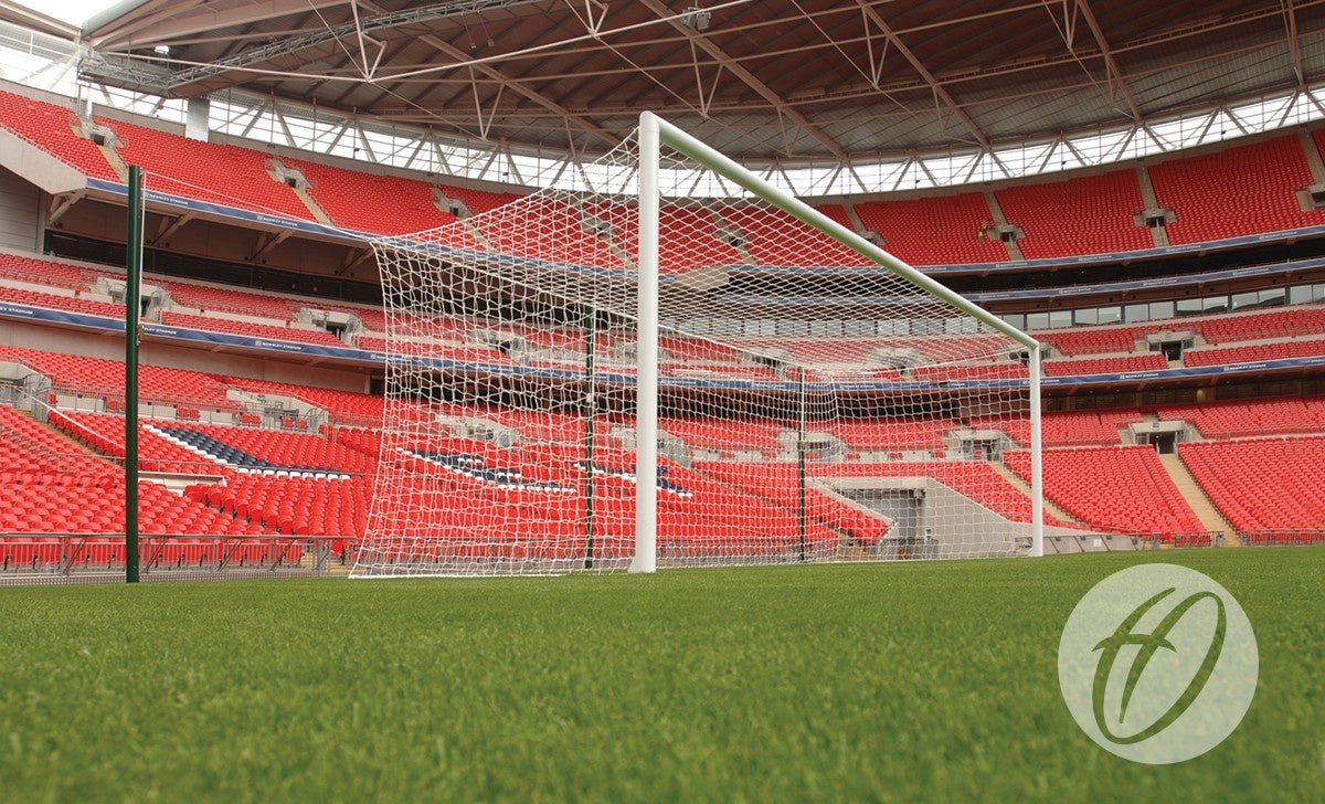 A close-up view of a soccer goal featuring the Harrod Sport Freehanging Net Support System - 6 Poles on a green field inside an empty stadium with red seats and a partially covered roof.