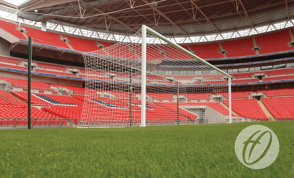 A close-up view of a soccer goal featuring the Harrod Sport Freehanging Net Support System - 6 Poles on a green field inside an empty stadium with red seats and a partially covered roof.