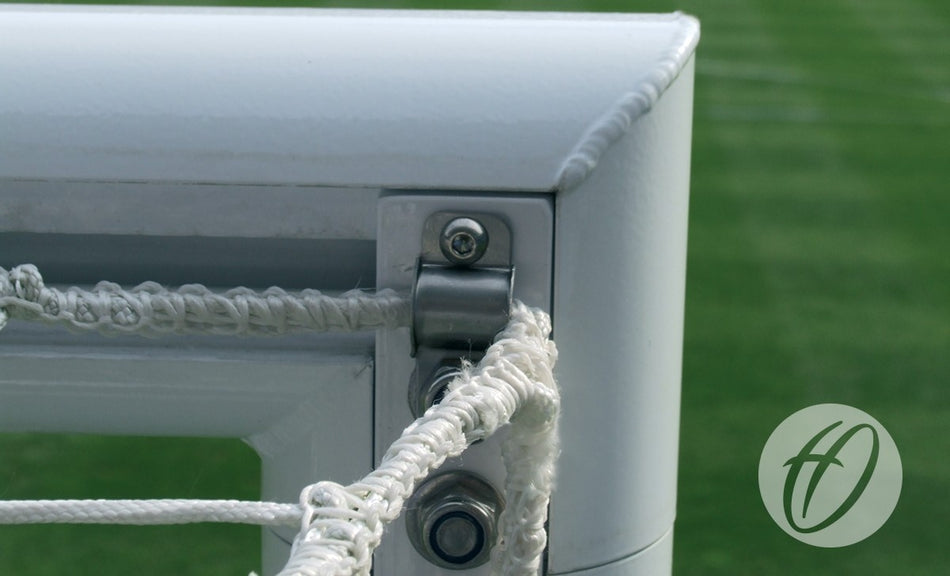 Close-up of a corner of the Harrod Sport 4G Stadium Goal - Junior, highlighting the aluminium frame, metal fixtures, and white netting against a background of blurred green artificial turf. The lower right corner features a circular logo with an "f".