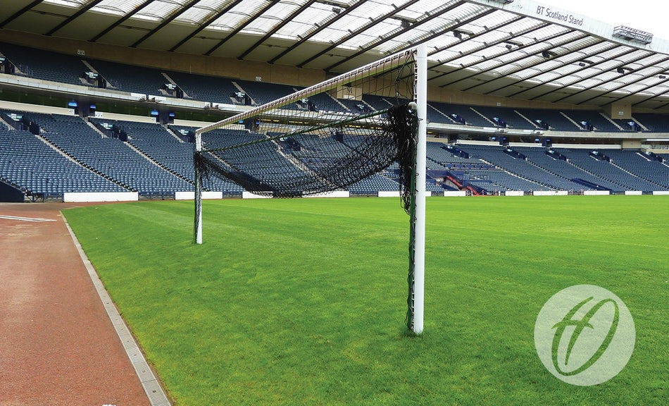 A well-lit soccer stadium with blue seats and a green field features the Harrod Sport 4G Stadium Goal - Senior with a black net. The venue has a partially covered roof and a logo on the grass in the foreground, evoking the style of Wembley Stadium.