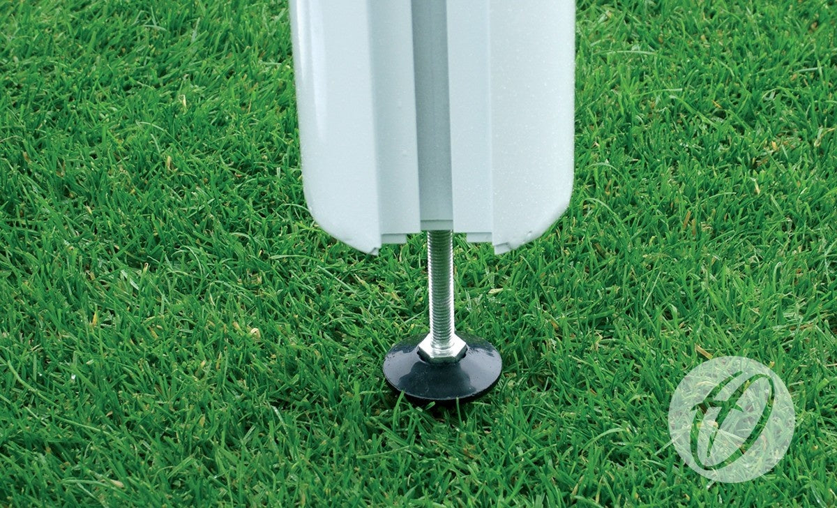 Close-up of the metal base of a Harrod Sport 4G Stadium Goal - Senior, securely anchored in green grass, with part of the white goal structure visible above. A translucent stylized “F” logo appears in the bottom right corner.