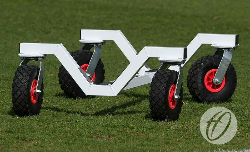 A set of Harrod Sport Portagoal Trolleys, white with a zigzag frame, four large black wheels and red hubs, is positioned on green grass. A circular logo with stylized letters appears in the lower right corner.