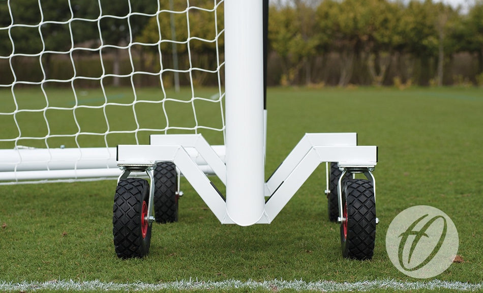 Close-up of Harrod Sport Portagoal Trolleys attached to the base of a white soccer goal, showing sturdy wheels for easy movement across a grassy field with trees and the brand logo in the background.