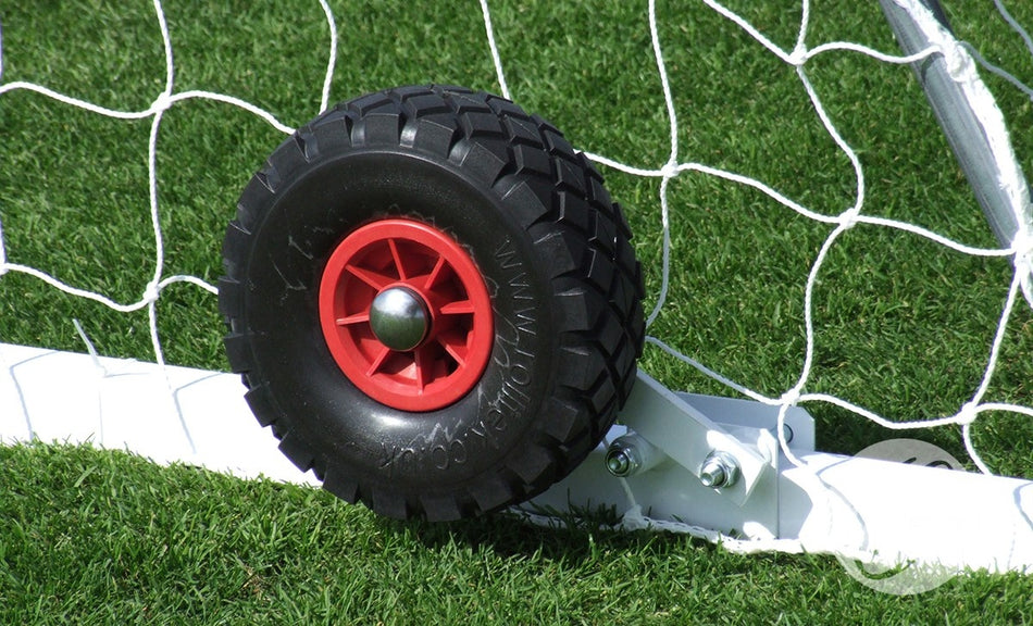 A close-up of a Harrod Sport Flip-over Wheel for Freestanding Steel Goals, featuring a black puncture proof tire and red hub attached to the soccer goal frame on green grass, with the white net in the background.