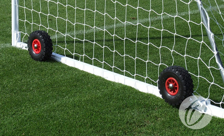 A close-up of a Harrod Sport freestanding steel goal with Flip-over Wheels, featuring two black and red puncture proof wheels at the base of the white metal frame and net, standing on green grass.