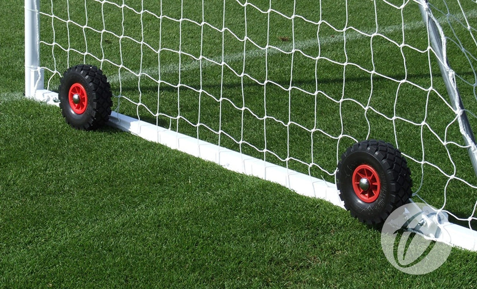 A close-up of a Harrod Sport freestanding steel goal with Flip-over Wheels, featuring two black and red puncture proof wheels at the base of the white metal frame and net, standing on green grass.