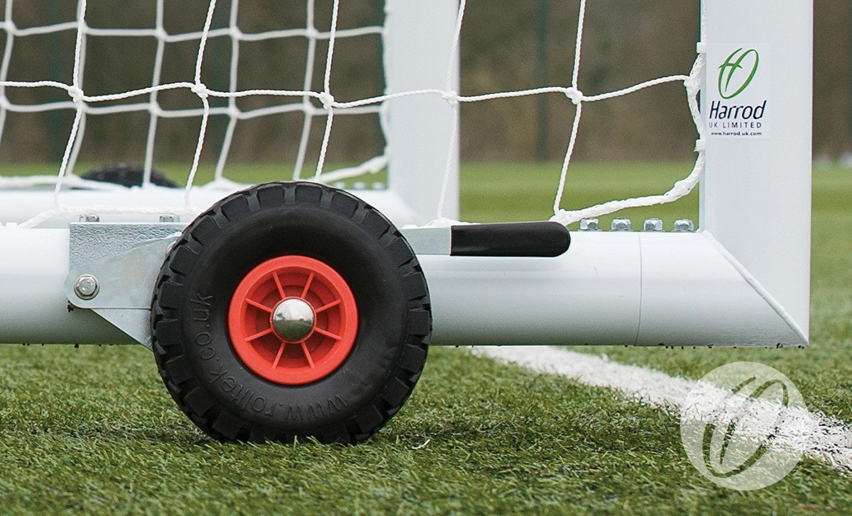Close-up of a white metal soccer goalpost with netting on artificial turf, featuring a Harrod Sport Set of 8 Hi-raise Football Goal Wheels—black and red, puncture-proof—attached to the frame with visible branding.