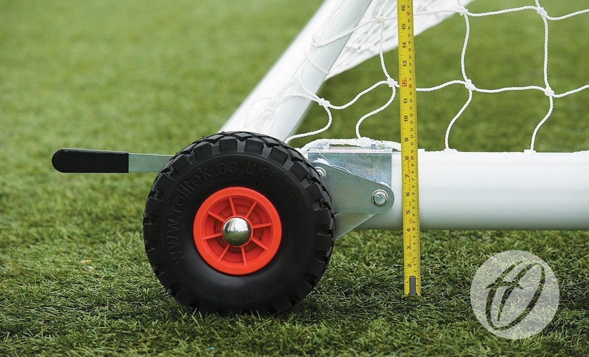 A close-up of a Harrod Sport Set of 8 Hi-raise Football Goal Wheels shows a black wheel with red hub attached to the soccer goal base on grass, and a yellow measuring tape standing upright next to the white frame.