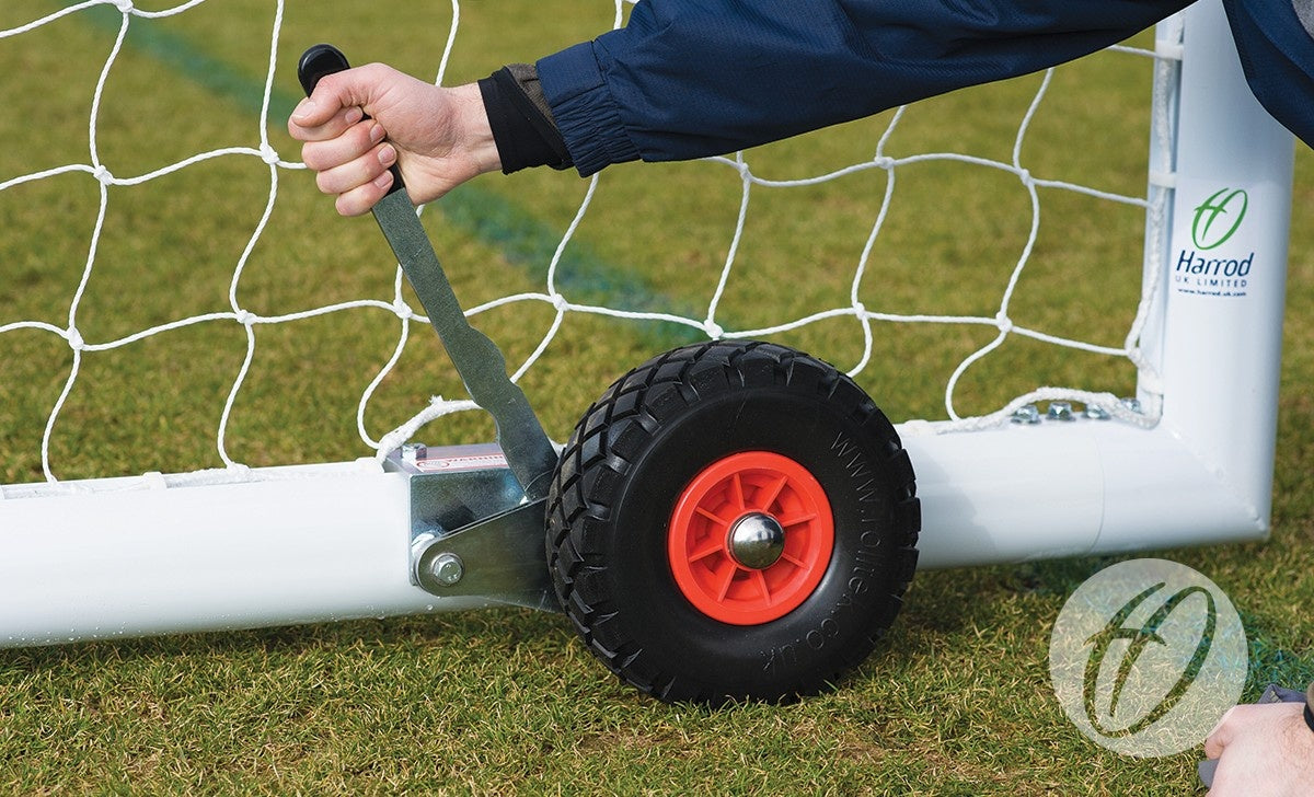 A person uses a metal lever to adjust a black and red puncture-proof wheel from the Set of 8 Hi-raise Football Goal Wheels by Harrod Sport, attached to a white soccer goalpost on grass. The Harrod Sport logo appears on the post.