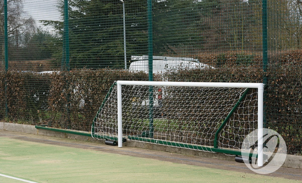 A Harrod Sport 4G Fence Folding Goal (3.66m x 1.22m, 3.5m to 5m projection) is set up on an outdoor field near a fence and hedge, with parked cars and trees visible in the background.