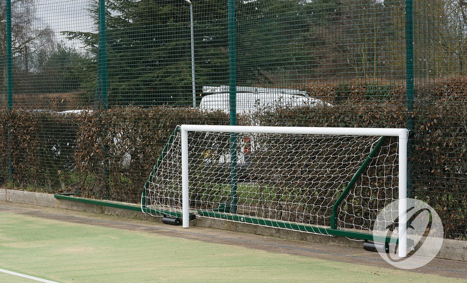 A Harrod Sport 4G Fence Folding Goal (3.66m x 1.22m, 3.5m to 5m projection) is set up on an outdoor field near a fence and hedge, with parked cars and trees visible in the background.