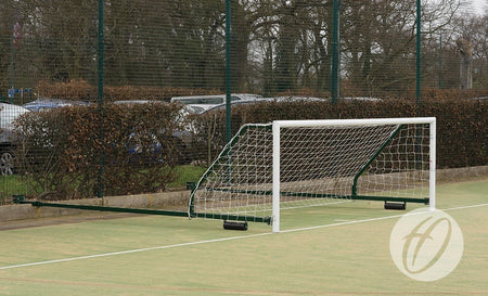 A Harrod Sport 4G Fence Folding Goal (3.66M x 1.22M, 3.5M to 5M projection) with its net attached is lying on its side in an outdoor field, with trees, bushes, parked cars, and a wire fence visible in the background.