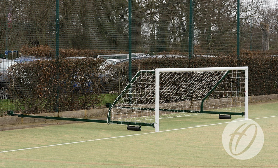 A Harrod Sport 4G Fence Folding Goal (3.66M x 1.22M, 3.5M to 5M projection) with its net attached is lying on its side in an outdoor field, with trees, bushes, parked cars, and a wire fence visible in the background.