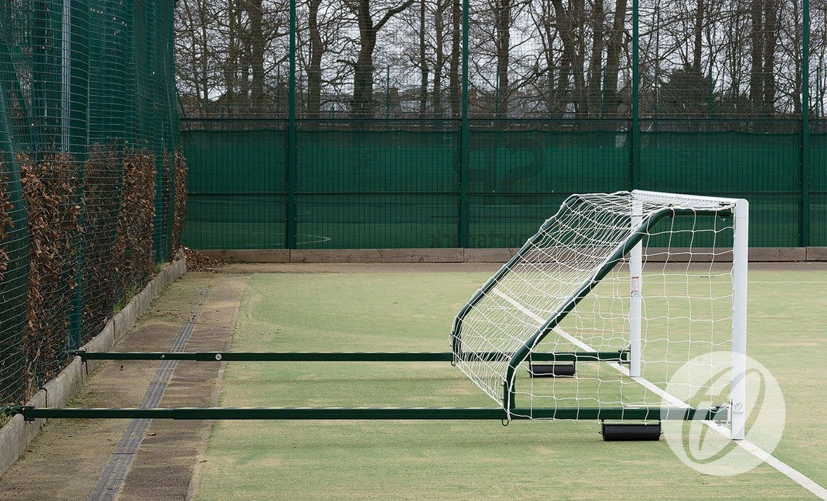 A Harrod Sport 4G Fence Folding Goal (3.66m x 1.22m, 3.5m to 5m projection) with wheels is lying on its side on an outdoor field next to a fenced area with trees in the background. This portable, folding 5-a-side goal is ideal for use on synthetic surface football pitches.